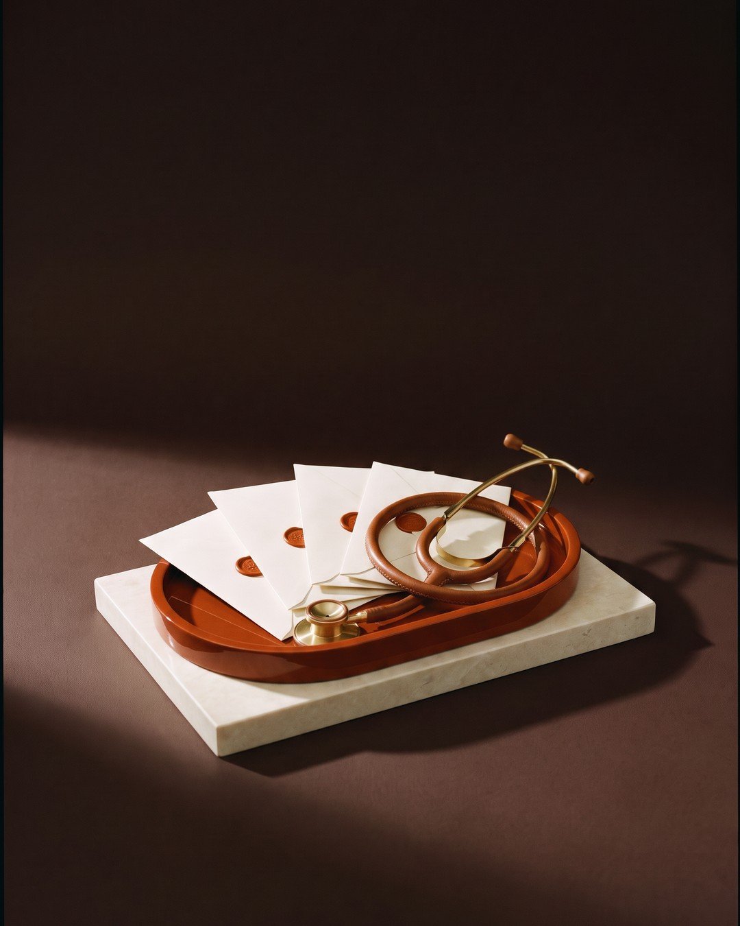 Four sealed ivory envelopes and a leather-and-brass stethoscope on a lacquered tray, against deep espresso brown